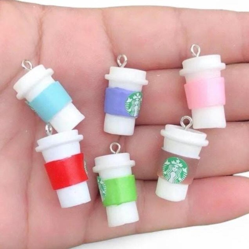 May include: Miniature coffee cup charms in white, with colorful bands in blue, purple, pink, red, and green. Each charm features a green logo and a silver loop for attaching to jewelry or crafts. The charms are held in a hand.