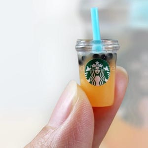 Pu&ograve; includere: Miniatura di bubble tea con una cannuccia blu. La tazza trasparente contiene un liquido giallo e perle di tapioca marrone scuro. Un logo verde e bianco &egrave; visibile sulla tazza.