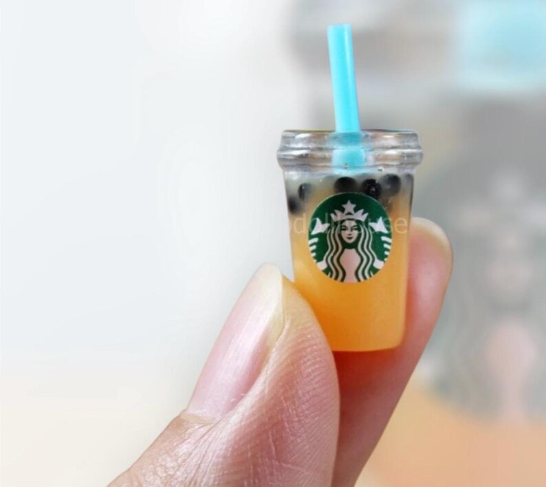 Pu&ograve; includere: Miniatura di bubble tea con una cannuccia blu. La tazza trasparente contiene un liquido giallo e perle di tapioca marrone scuro. Un logo verde e bianco &egrave; visibile sulla tazza.