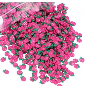 May include: A pile of small, pink strawberry-shaped polymer clay pieces with white dots and green leaves. The clay pieces are scattered, with some appearing to be in a clear plastic bag. The strawberries are a vibrant pink color.