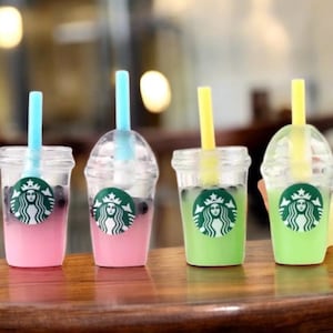 Pu&ograve; includere: Quattro mini bicchieri di plastica trasparente con bevande colorate e cannucce. Le bevande sono rosa e verdi, con un logo Starbucks sul davanti. Ogni bicchiere ha una cannuccia di colore diverso: blu e giallo.
