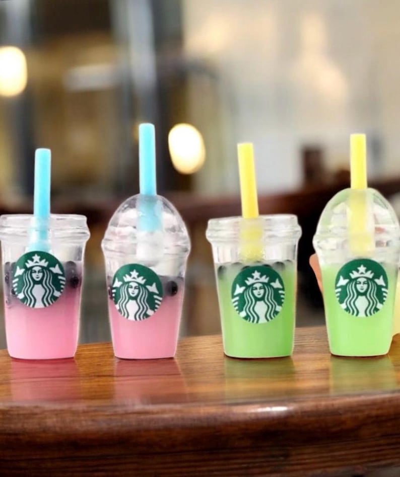 Pu&ograve; includere: Quattro mini bicchieri di plastica trasparente con bevande colorate e cannucce. Le bevande sono rosa e verdi, con un logo Starbucks sul davanti. Ogni bicchiere ha una cannuccia di colore diverso: blu e giallo.