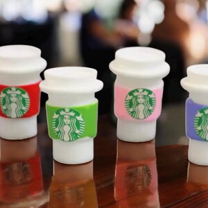 May include: Four white miniature coffee cups with white lids and colorful sleeves. Each cup has a green Starbucks logo on the sleeve. The sleeves are red, green, pink, and purple. The cups are on a brown surface.