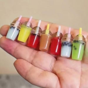Pu&ograve; includere: Miniature bevande al t&egrave; con le bolle in vari colori, tra cui giallo, rosso e verde, con bicchieri di plastica trasparente, cannucce e coperchi. Le bevande sono tenute in una mano, mostrando la variet&agrave; di gusti e le piccole dimensioni.