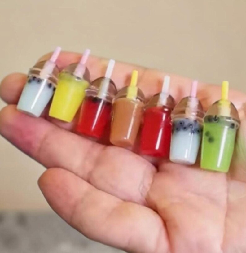 Pu&ograve; includere: Miniature bevande al t&egrave; con le bolle in vari colori, tra cui giallo, rosso e verde, con bicchieri di plastica trasparente, cannucce e coperchi. Le bevande sono tenute in una mano, mostrando la variet&agrave; di gusti e le piccole dimensioni.