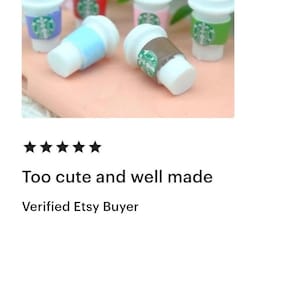 May include: Miniature coffee cups with white lids and colorful sleeves. The sleeves are in various colors, including blue, brown, green, and pink, each with a green logo. The cups are arranged on a light pink surface with green foliage in the background.