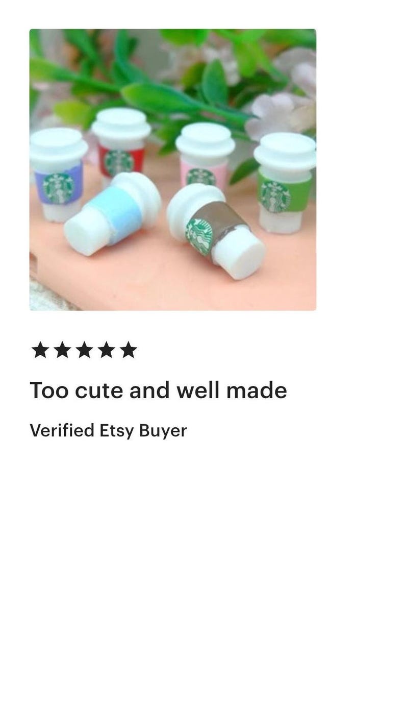 May include: Miniature coffee cups with white lids and colorful sleeves. The sleeves are in various colors, including blue, brown, green, and pink, each with a green logo. The cups are arranged on a light pink surface with green foliage in the background.