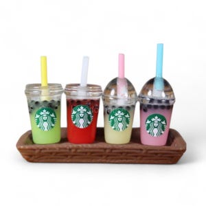 Pu&ograve; includere: Quattro miniature di bubble tea in bicchieri trasparenti con cannucce, presentate su un vassoio marrone. Le bevande sono verdi, rosse, gialle e rosa, ognuna con un logo Starbucks. Le cannucce sono gialle, bianche, rosa e blu.
