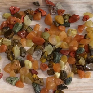 May include: A collection of colorful gemstone beads in shades of red, orange, yellow, green, and brown. The beads are of various shapes and sizes, some with holes for stringing. The gemstones appear to be polished and have a natural, earthy look.