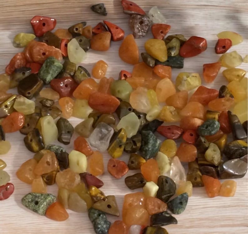 May include: A collection of colorful gemstone beads in shades of red, orange, yellow, green, and brown. The beads are of various shapes and sizes, some with holes for stringing. The gemstones appear to be polished and have a natural, earthy look.