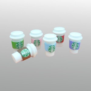 May include: Miniature coffee cups with white lids and colorful sleeves. The sleeves are green, pink, blue, red, and purple, each featuring a green logo. One cup is lying on its side, showcasing a brown sleeve.