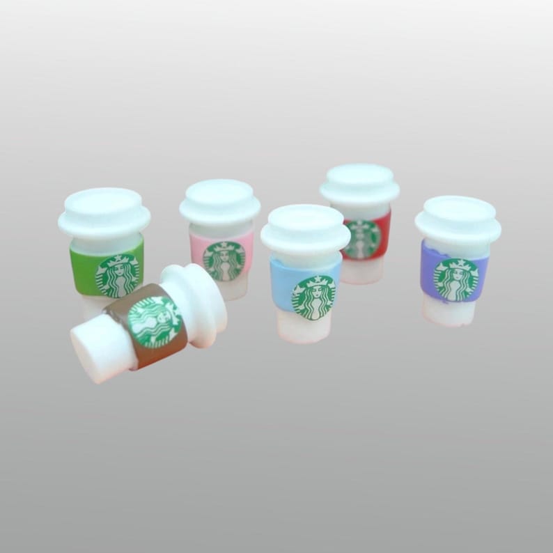 May include: Miniature coffee cups with white lids and colorful sleeves. The sleeves are green, pink, blue, red, and purple, each featuring a green logo. One cup is lying on its side, showcasing a brown sleeve.