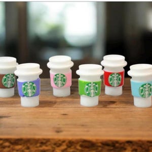 May include: Miniature white coffee cups with white lids and colorful sleeves. Each cup features the Starbucks logo in green. Sleeves are in brown, purple, pink, green, red, and blue. The cups are arranged on a wooden surface.