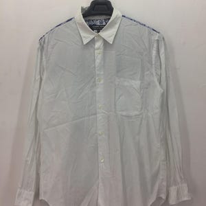 Puede incluir: Camisa blanca de manga larga con cuello cl&aacute;sico y bolsillo en el pecho. Los hombros y el interior del cuello presentan un estampado floral azul y blanco. La camisa est&aacute; colgada de una percha negra.