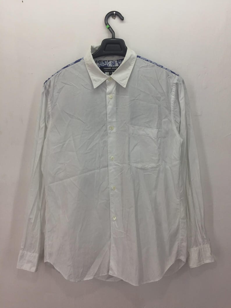 Puede incluir: Camisa blanca de manga larga con cuello cl&aacute;sico y bolsillo en el pecho. Los hombros y el interior del cuello presentan un estampado floral azul y blanco. La camisa est&aacute; colgada de una percha negra.