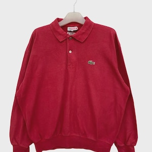 Può includere: Una polo rossa a maniche lunghe con colletto button-down e un piccolo logo verde a forma di coccodrillo sul petto. La camicia è appesa a un'appendiabiti su sfondo bianco. La camicia ha un aspetto vintage.