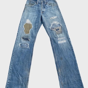 Puede incluir: Vaqueros de mezclilla azul claro con detalles desgastados, incluyendo rasgaduras y parches. Los vaqueros tienen bragueta de botones y un aspecto descolorido. Los vaqueros están extendidos, mostrando su corte de pierna recta. La etiqueta dice "P656".