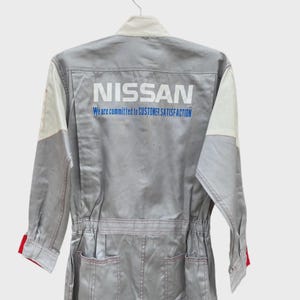 May include: A gray Nissan mechanic's jumpsuit with white and red accents. The back of the jumpsuit features the Nissan logo and the text "We are committed to CUSTOMER SATISFACTION". The jumpsuit has pockets and red stitching.