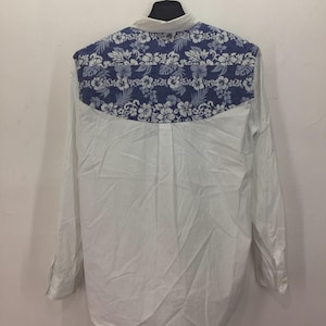 Puede incluir: Camisa blanca de manga larga con un estampado floral azul en la parte superior de la espalda. El dise&ntilde;o floral presenta flores y hojas blancas sobre un fondo azul. La camisa est&aacute; colgada en una percha negra.
