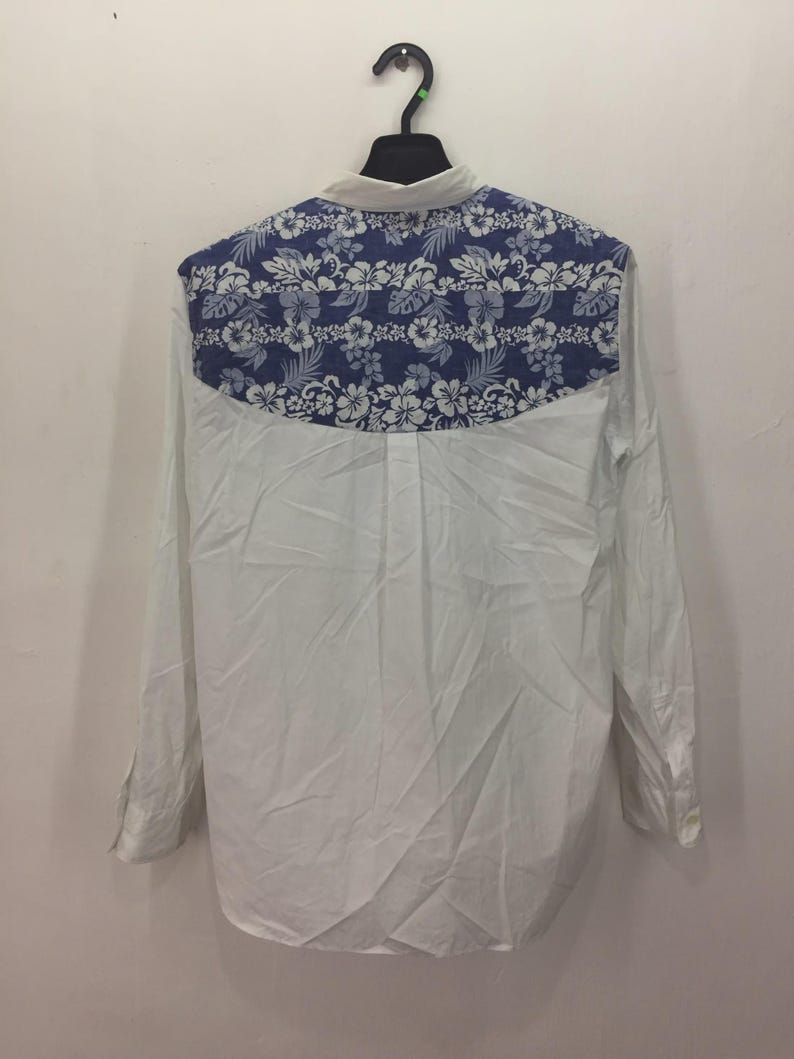 Puede incluir: Camisa blanca de manga larga con un estampado floral azul en la parte superior de la espalda. El dise&ntilde;o floral presenta flores y hojas blancas sobre un fondo azul. La camisa est&aacute; colgada en una percha negra.