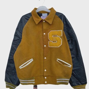 Könnte beinhalten: Senfgelbe Collegejacke mit schwarzen Ärmeln. Sie verfügt über einen großen weißen und gelben "S"-Aufnäher und weiße Akzente an den Taschen und Manschetten. Die Jacke hat eine Knopfleiste und einen gerippten Saum. Das Etikett lautet "W127 EMECAS".