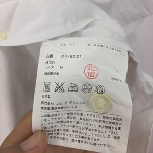 Puede incluir: Camisa de algod&oacute;n blanca con una etiqueta que muestra las instrucciones de cuidado y el texto "DK-B021". La etiqueta tambi&eacute;n incluye la talla "M" y los textos "綿100%" y "日本製".