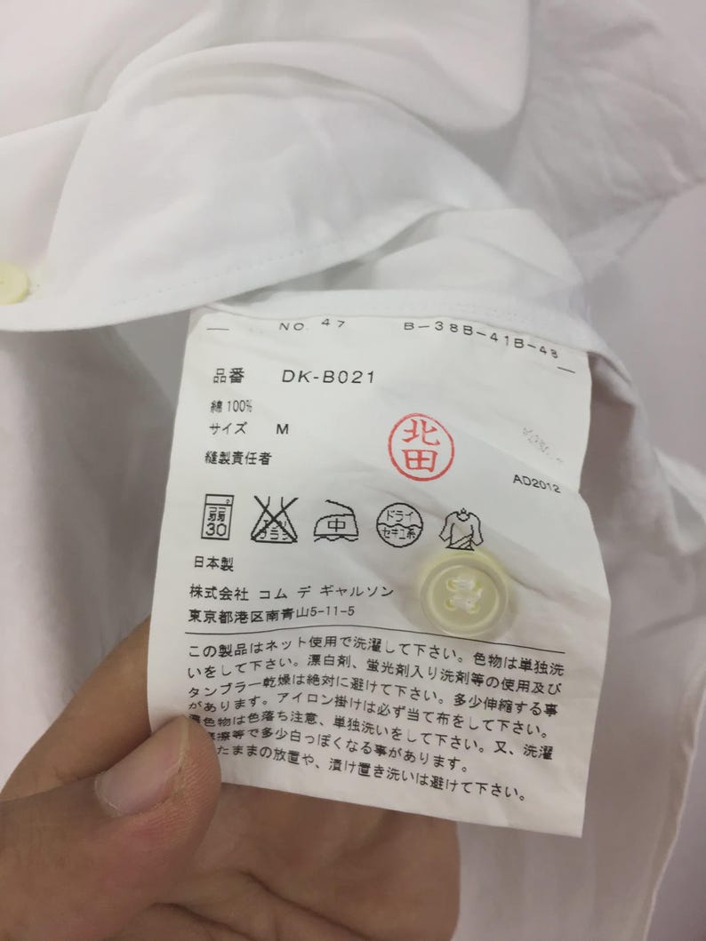 Puede incluir: Camisa de algod&oacute;n blanca con una etiqueta que muestra las instrucciones de cuidado y el texto "DK-B021". La etiqueta tambi&eacute;n incluye la talla "M" y los textos "綿100%" y "日本製".