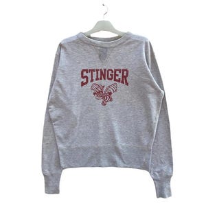 Può includere: Una felpa grigia mélange con la scritta "STINGER" in lettere bordeaux e una grafica a forma di ape. La parte superiore a maniche lunghe ha girocollo e polsini e orlo a coste.