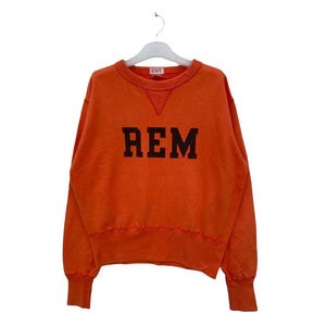 Può includere: Felpa arancione con la scritta "REM" a blocchi neri sul petto. Ha girocollo, polsini e orlo a coste. L'indumento è appeso a una gruccia bianca. Stile casual.