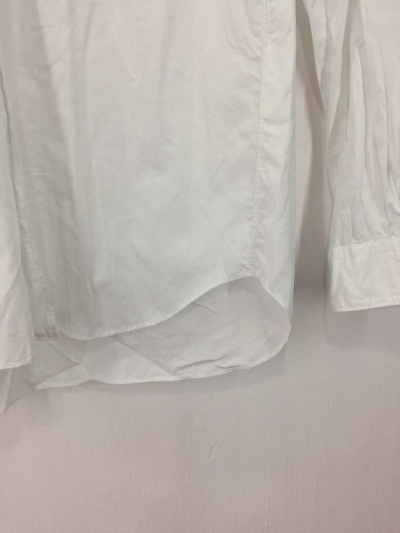 Puede incluir: Primer plano de una camisa blanca abotonada. La camisa est&aacute; hecha de una tela ligera y presenta mangas largas con pu&ntilde;os abotonados. El dobladillo inferior es curvo.