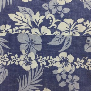 Puede incluir: Primer plano de una tela azul con un estampado floral blanco. El dise&ntilde;o presenta grandes flores de hibisco, flores m&aacute;s peque&ntilde;as y hojas estilizadas. La tela parece ser de algod&oacute;n o similar, con una superficie ligeramente texturizada.