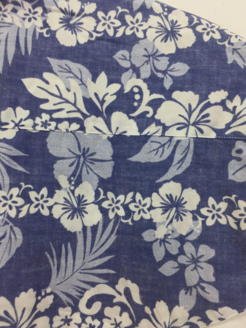 Puede incluir: Primer plano de una tela azul con un estampado floral blanco. El dise&ntilde;o presenta grandes flores de hibisco, flores m&aacute;s peque&ntilde;as y hojas estilizadas. La tela parece ser de algod&oacute;n o similar, con una superficie ligeramente texturizada.