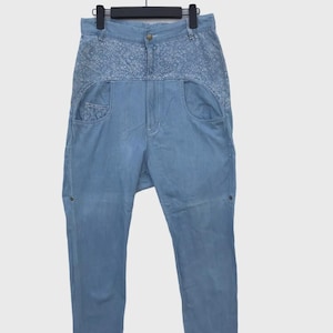 Może przedstawiać: Jasnoniebieskie dżinsy z ozdobnym haftowanym panelem w talii i kieszeniami. Spodnie wiszą na czarnym wieszaku. Spodnie mają luźny krój i zapięcie na guziki.