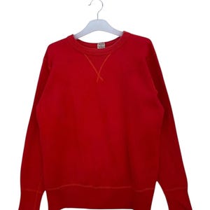 Può includere: Una felpa rossa tinta unita con girocollo e maniche lunghe, appesa a una gruccia bianca. Le cuciture intorno allo scollo sono visibili. Lo sfondo è bianco.