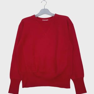 Può includere: Una felpa rossa tinta unita con girocollo e maniche lunghe. I polsini e l'orlo sono a coste. La felpa è appesa a una gruccia grigia su sfondo bianco.