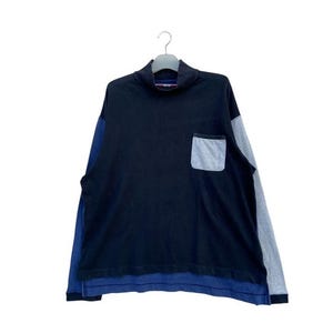 Può includere: Una maglia a maniche lunghe con collo alto e tasca. La maglia è principalmente blu navy con una tasca grigia e maniche grigie e blu. L'orlo inferiore è blu.