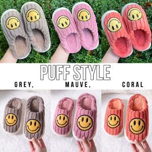 May include: Three pairs of fuzzy slippers with a smiley face design. The slippers are in grey, mauve, and coral.
