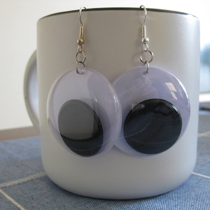 May include: A pair of earrings with clear plastic circles and black swirl designs. The earrings are hanging from a white mug.