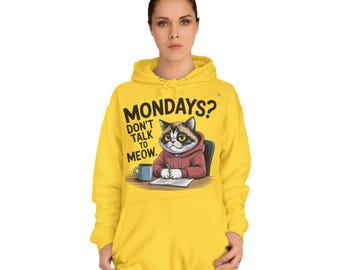 Puede incluir: Sudadera con capucha amarilla con un gráfico de un gato gruñón con una sudadera roja, sentado en un escritorio con una taza y un bloc de notas. El texto en la sudadera dice "MONDAYS? DON'T TALK TO MEOW."