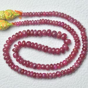 May include: A strand of small, round, deep pink ruby beads arranged in a loose spiral. The beads are uniform in shape and size, featuring a polished finish. At one end of the strand, there are two decorative, textured gold-colored tassels.