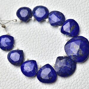 May include: A strand of blue lapis lazuli gemstone beads with gold flecks. The beads are faceted and have a teardrop shape. The beads are strung on a white thread.