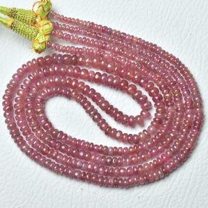 Op de afbeelding: Slierten kleine, ronde, roze edelstenen, mogelijk robijnen, zijn in een vloeiend patroon gerangschikt. De kralen hebben een consistente grootte en kleur, met een lichte variatie in toon. De strengen zijn samengebonden met een groene en gouden kwast.