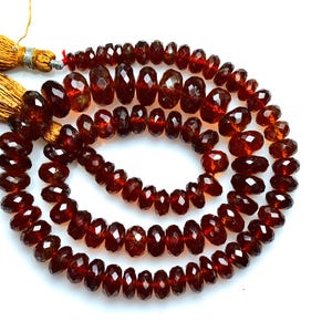 May include: A strand of deep red, faceted, circular garnet beads arranged in a spiral pattern. The strand is finished with two golden-brown silk tassels attached at the top, featuring metallic silver-colored thread bindings at the base of the tassels.