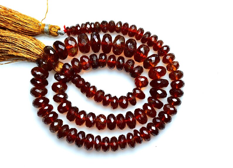 May include: A strand of deep red, faceted, circular garnet beads arranged in a spiral pattern. The strand is finished with two golden-brown silk tassels attached at the top, featuring metallic silver-colored thread bindings at the base of the tassels.