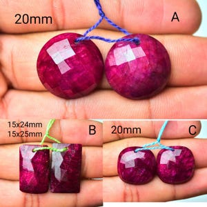 Op de afbeelding: Close-up van drie sets gefacetteerde, dieprode edelstenen. De eerste set heeft twee ronde stenen, elk met een diameter van 20 mm, geregen op blauwe draad. De tweede set heeft twee rechthoekige stenen, 15x24mm en 15x25mm, op groene draad. De derde set heeft twee vierkante stenen, elk 20 mm.
