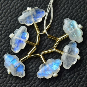 Puede incluir: Un colgante con forma de copo de nieve hecho con seis cuentas de piedra lunar iridiscente, cada una con forma de nube, conectadas por separadores de color dorado. El colgante está enhebrado en un hilo blanco.