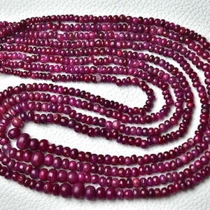 Könnte beinhalten: Eine Strähne aus Rubinperlen, etwa 3 mm groß, in tiefroter Farbe. Die Perlen sind auf einem durchsichtigen Faden aufgefädelt und in einem kreisförmigen Muster angeordnet.