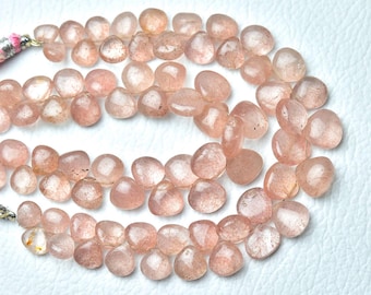 May include: Close-up of peach-coloured, teardrop-shaped gemstones. The beads are translucent, with slight colour and texture variations. The gemstones are strung together, possibly for a necklace or bracelet.