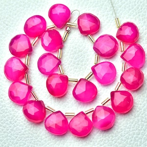 Könnte beinhalten: Stränge aus facettierten, leuchtend pinkfarbenen Edelsteinperlen. Jede herzförmige Perle hat einen goldfarbenen Metallakzent. Die Perlen sind aufgefädelt und bilden eine Halsketten- oder Armbandkomponente. Der Hintergrund ist hell und neutral.