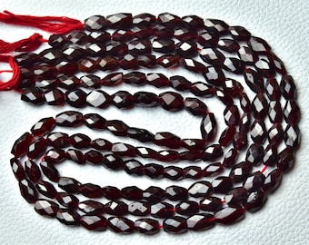 Puede incluir: Primer plano de varios hilos de cuentas de granate rojo oscuro. Las cuentas tienen forma ovalada con múltiples facetas, ensartadas en hilo rojo. Las cuentas son de un color rojo intenso y las facetas reflejan la luz, creando un efecto brillante. Cada cuenta mide aproximadamente 0,8 cm.